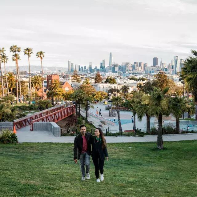 A couple walks toward the camera with Dolores Park 和 San Francisco Skyline behind them.